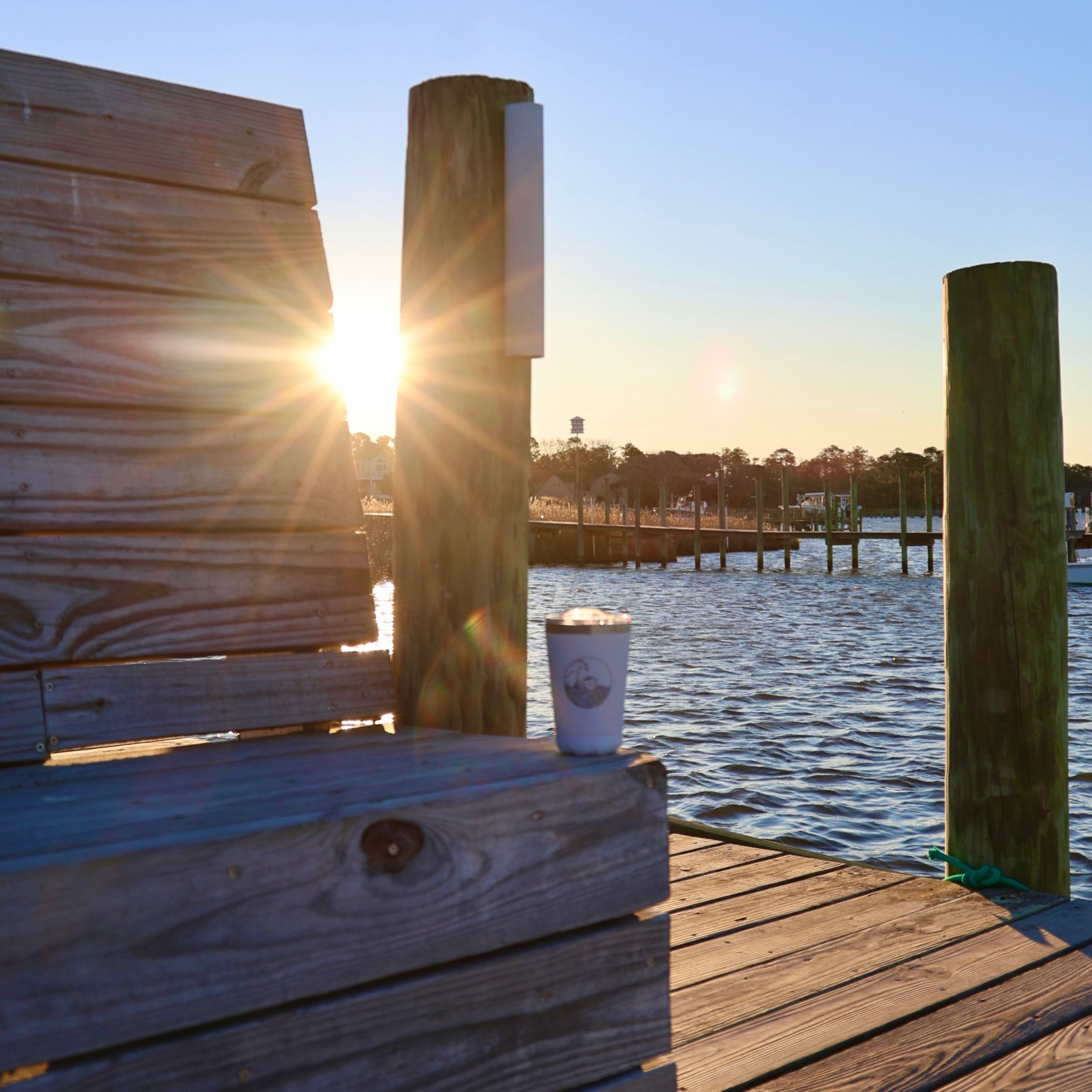 A wooden dock with a bench and a cup, overlooking a body of water with a pier in the background.