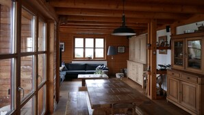 Ruim houten interieur van een blokhut met een eethoek, keuken en een groot raam voor natuurlijk licht.