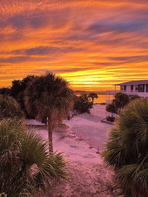 A beachfront property with a sandy pathway leading to a white building with a balcony, surrounded by palm trees and a sunset sky. - Spacious House, 1 Back, Beach Access, Big Porches, Inlet Views, Pretty Sunsets! (Tybee Island)