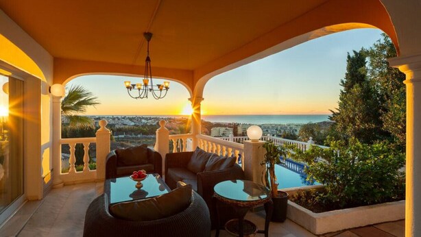 A balcony with a sofa, coffee table, and potted plant, overlooking a cityscape and a body of water.