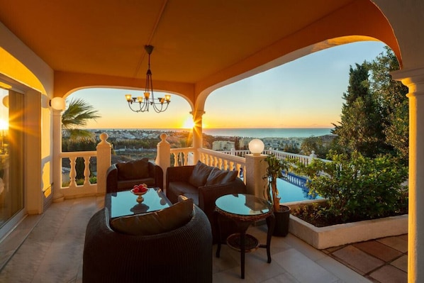 A balcony with a sofa, coffee table, and potted plant, overlooking a cityscape and a body of water.