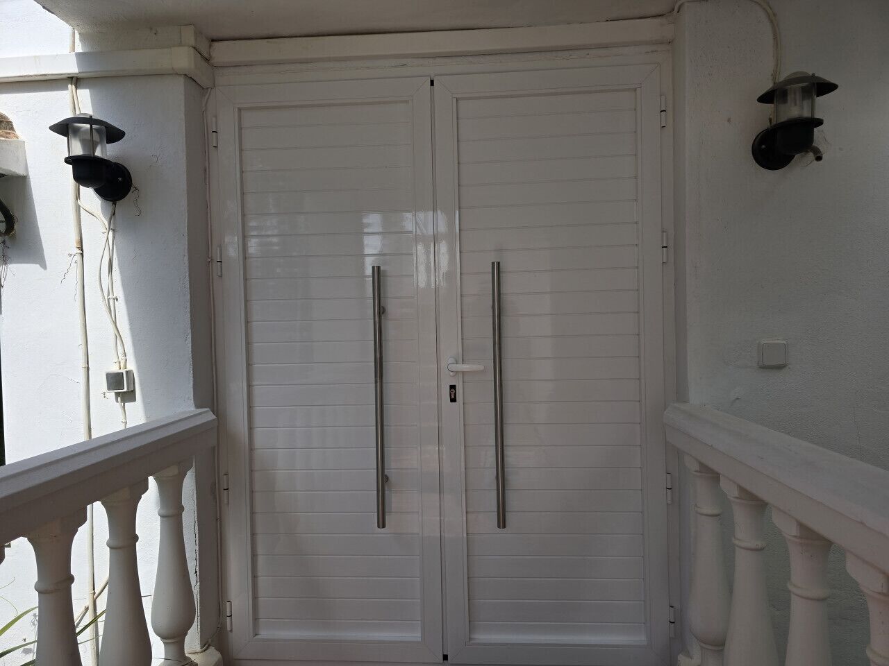 A white door with two handles, a white railing, and a wall-mounted light fixture.