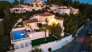 A large, multi-story house with a swimming pool, surrounded by lush greenery and palm trees.