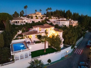 A large, multi-story house with a swimming pool, surrounded by lush greenery and palm trees.