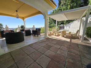En uteplats med ett kaklat golv, vita pelare och en pergola som ger skugga.