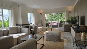 Un salón amplio con un sofá grande, una mesa de centro y un televisor. Hay un balcón con vistas a zonas verdes.