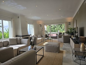 A spacious living room with a large sofa, coffee table, and a TV. There is a balcony with a view of greenery. - Luxury Villa with Heated Pool, Stunning Sea Views & Near Beach (Las Lagunas de Mijas)