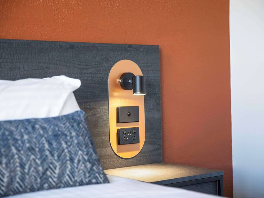 Habitación de hotel moderna con cabecera de madera, mesita de noche y panel de control en la pared.