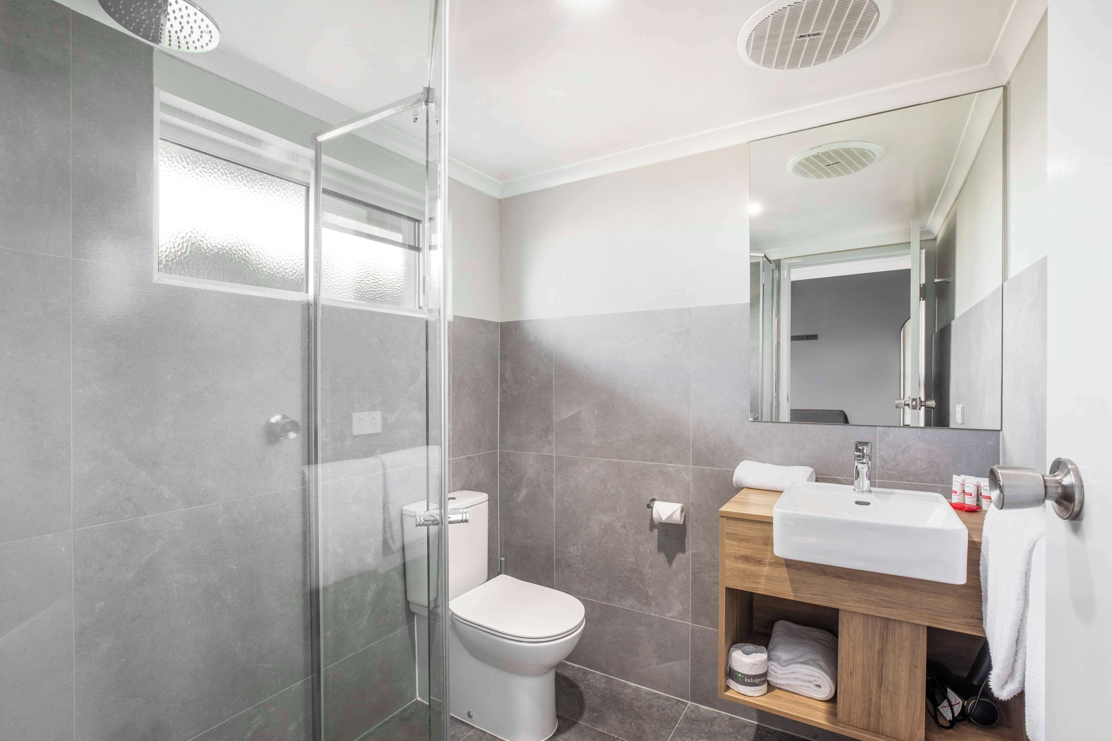 A modern bathroom with a glass shower enclosure, a white toilet, a wooden vanity with a sink, and a large mirror.