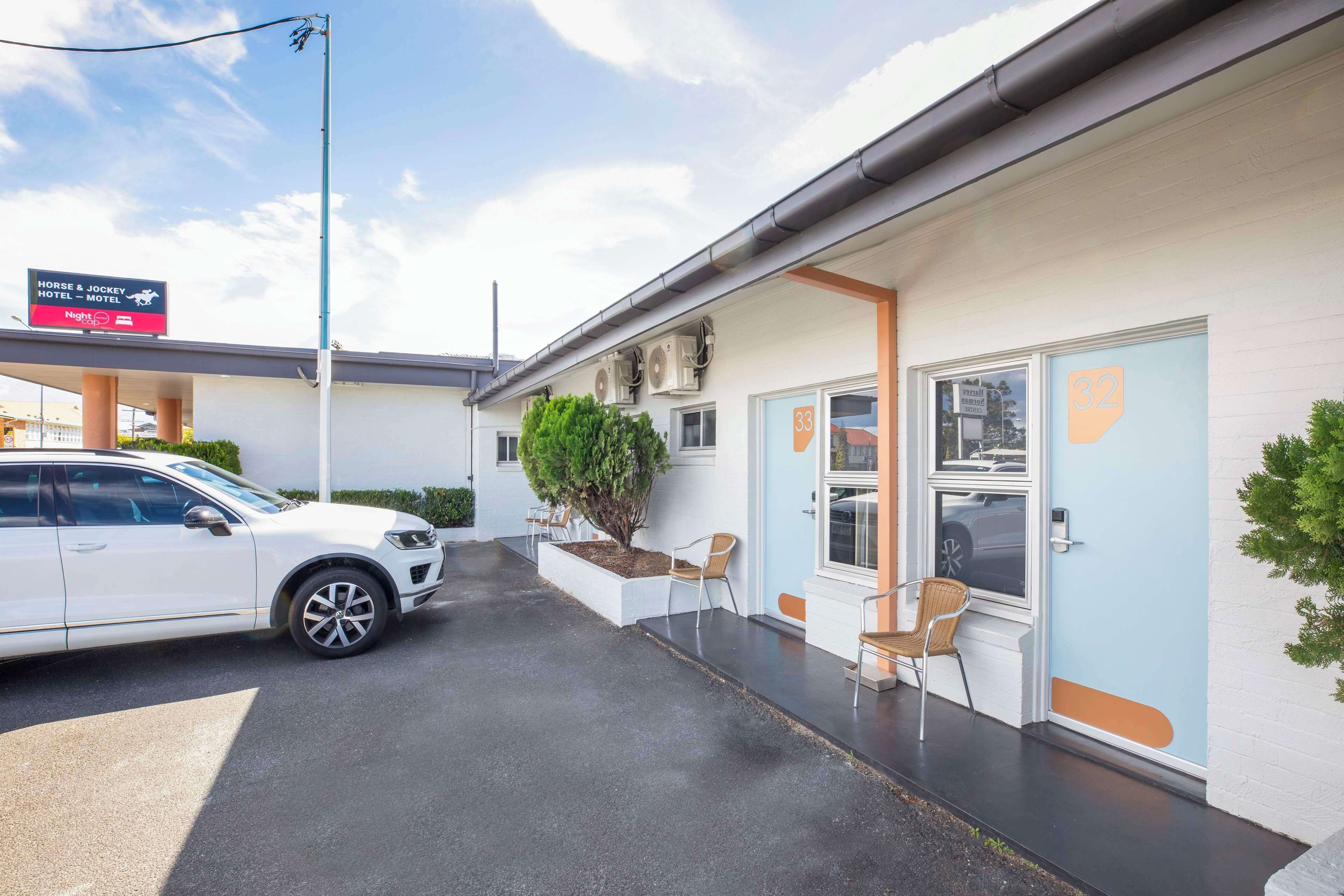 L’entrée d’un hôtel, avec une voiture garée devant, une enseigne au-dessus et un petit jardin agrémenté de chaises.