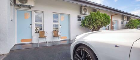 Una hilera de puertas numeradas, unidades de aire acondicionado y un coche blanco estacionado frente a un edificio.