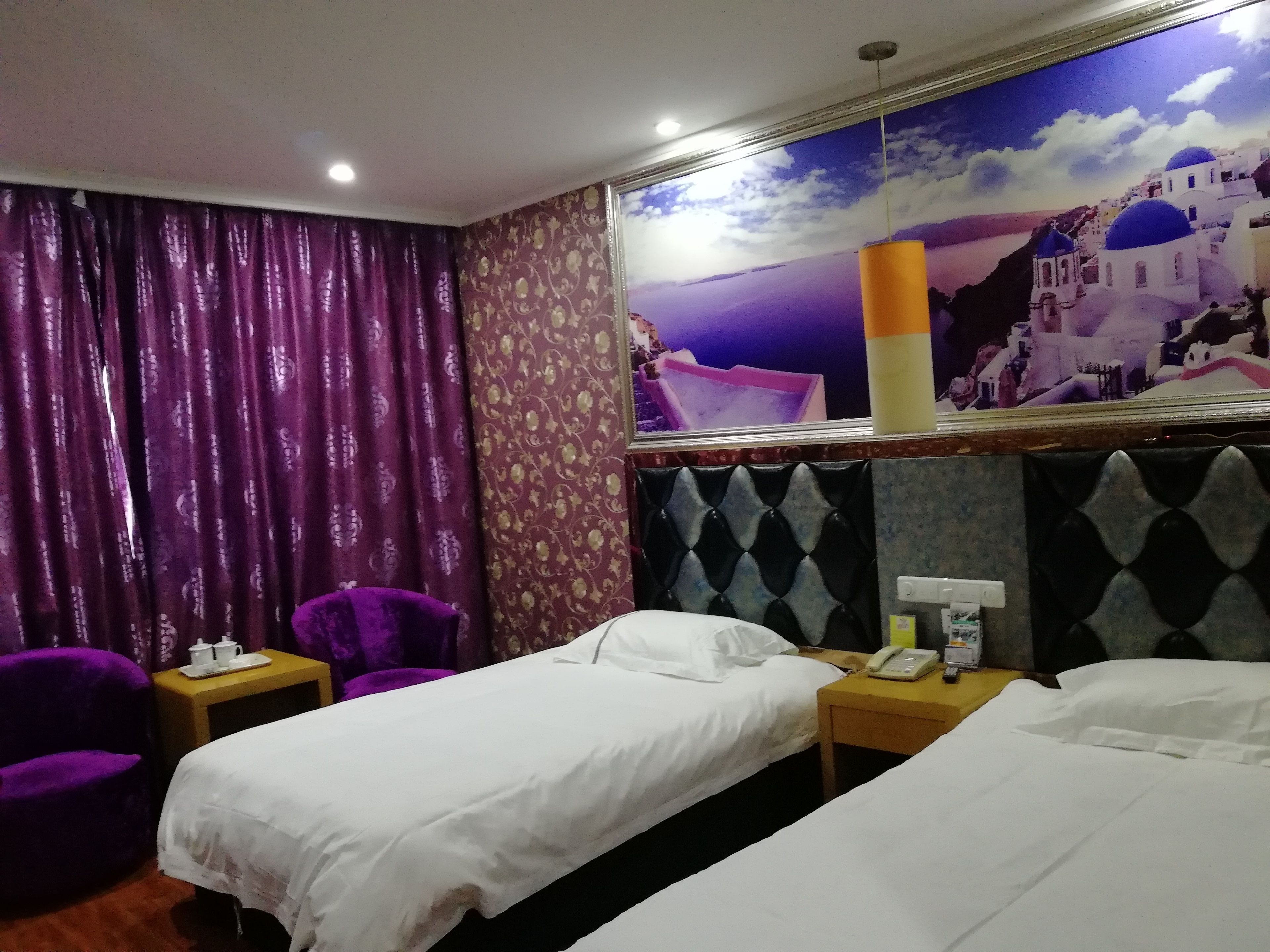 Une chambre d’hôtel avec deux lits simples, un fauteuil violet, une petite table avec un téléphone et une tasse, et une fresque murale représentant un paysage côtier.