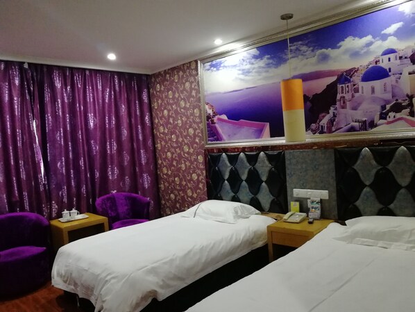 Una habitación de hotel con dos camas individuales, un sillón morado, una mesita con un teléfono y una taza, y un mural en la pared que representa una escena costera.