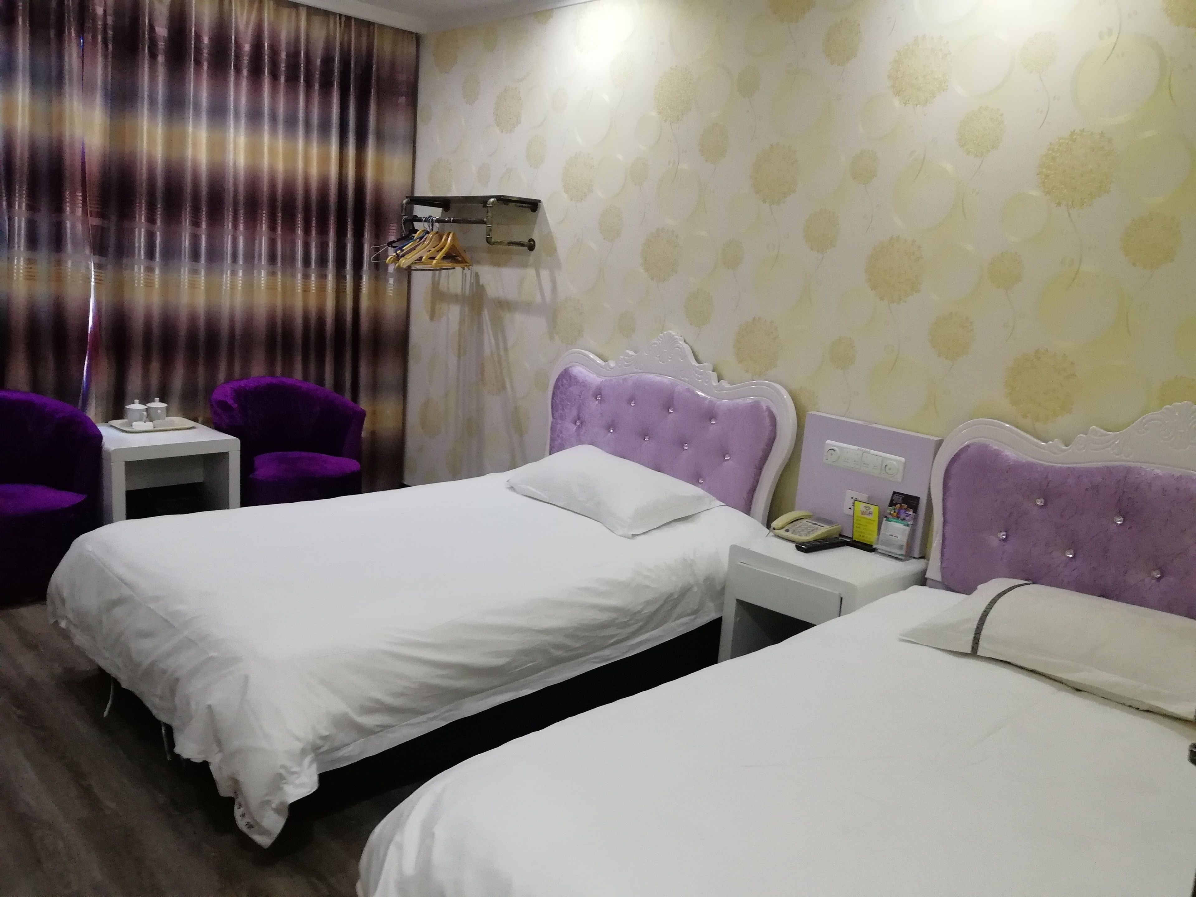 Hotellihuone, jossa on kaksi sänkyä, pieni pöytä, violetit tuolit ja seinälle kiinnitetty hylly, jossa on hygieniatarvikkeita.