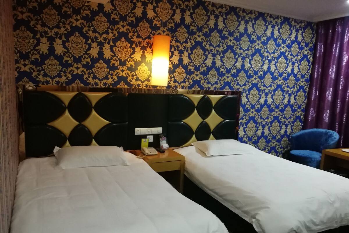 Une chambre d’hôtel avec deux lits, une tête de lit ornée d’un motif géométrique, une table de chevet avec un téléphone et une lampe, et une chaise bleue.