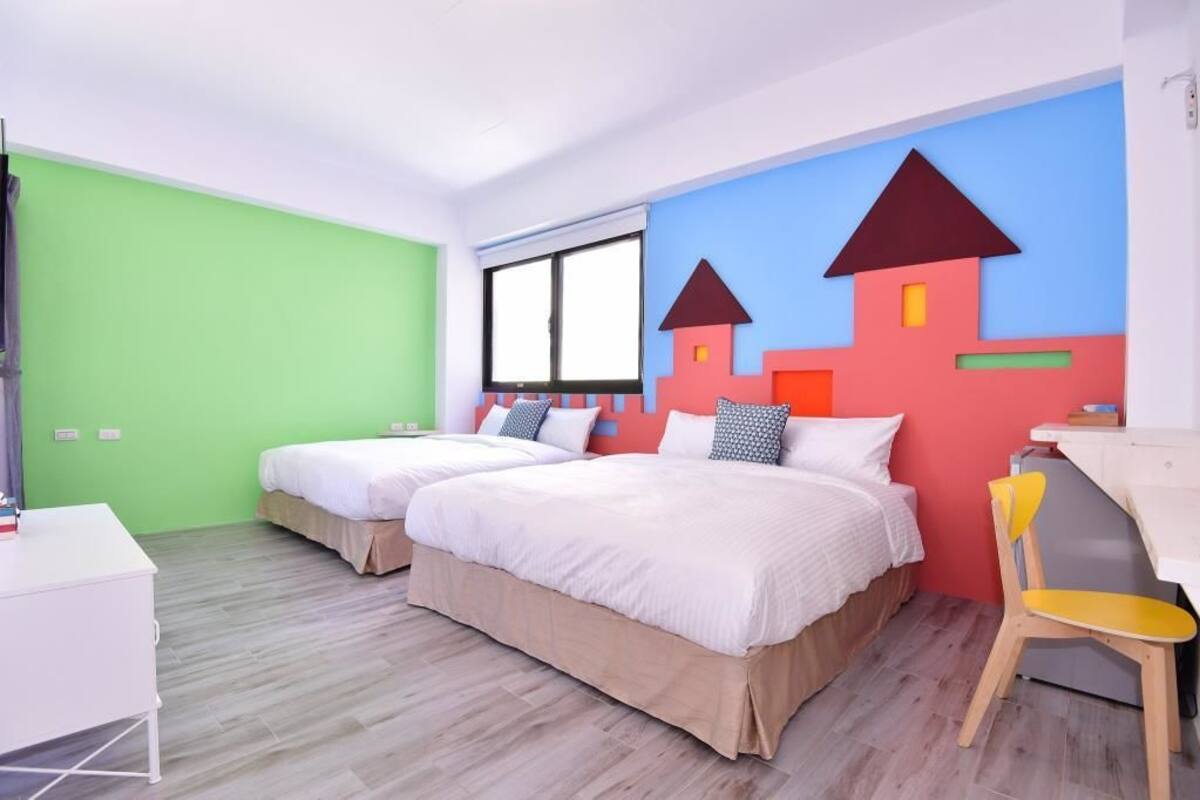 Habitación de hotel con dos camas, un escritorio y una silla. Cuenta con una ventana con persianas, una pared verde y una decoración mural colorida.