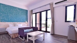 Un dormitorio con una cama, un sillón morado, una mesa blanca y un ventanal con cortinas.