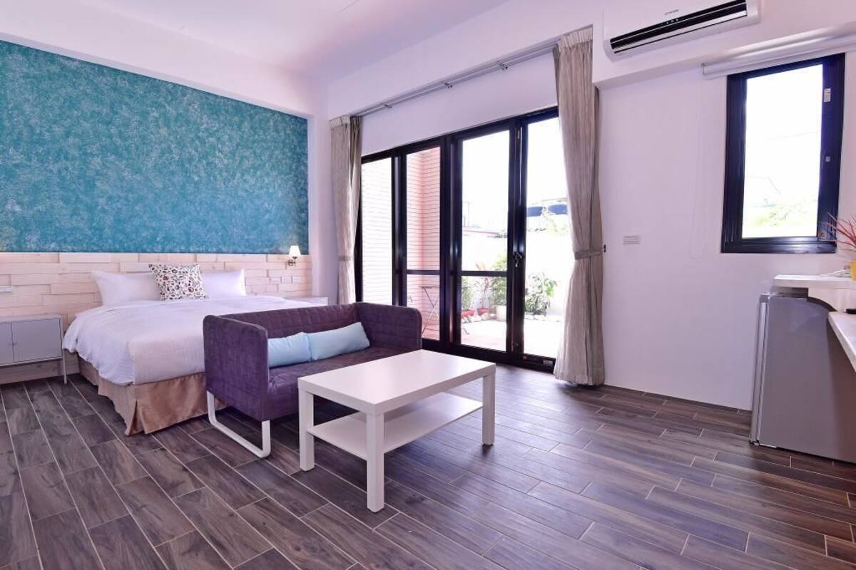 Un dormitorio con una cama, un sillón morado, una mesa blanca y un ventanal con cortinas.