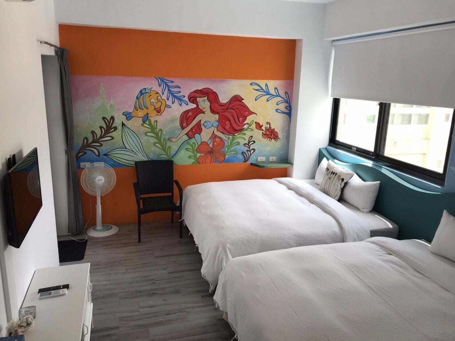 A hotel room with two beds, a mural on the wall, a desk with a chair, and a window with a view.