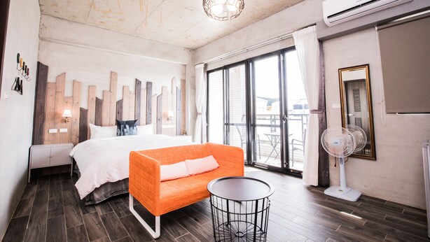 Una habitación de hotel moderna con una cama, un sofá naranja, una pequeña mesa redonda, un ventilador de pie y un ventanal con vistas.