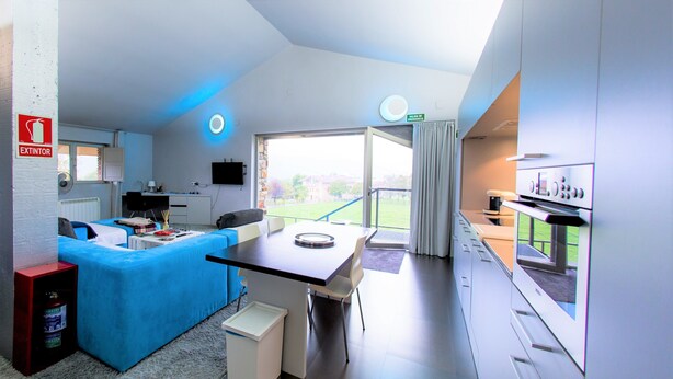 A modern hotel room with a blue sofa, a dining table, a kitchen area, and a view of the outdoors.