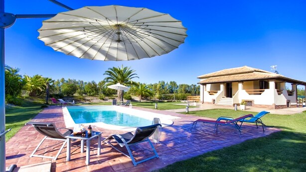 Un área junto a la piscina con sillones de descanso, una mesa y una sombrilla grande.
