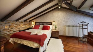 Een slaapkamer met schuine plafonds, een stenen muur, houten balken, een bed met rode beddengoed, bedlampen, een radiator en een klein houten tafeltje.