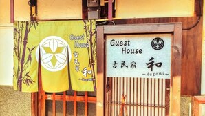 Ein traditionelles japanisches Gebäude mit hölzernem Eingang, einem Schild mit der Aufschrift 'Guest House' und 'Nagomi', verfasst in Englisch und Japanisch, sowie einem dekorativen Bambusmuster.