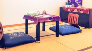 Ein Zimmer mit Tatami-Boden, einem violetten Tisch mit Uhr und Büchern sowie zwei blauen Kissen.