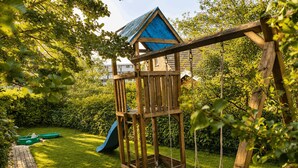 A wooden play structure with a slide and swings in a lush garden. - Top vacation home,7 p.,sauna,2 baths,WLAN,fireplace,beach kitchen,terrace,dog,playground (Dornum)
