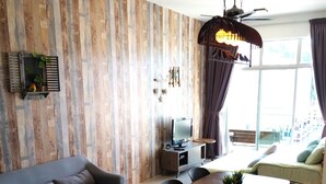A modern living room with a sofa, dining table, and chairs. There is a TV, a small desk, and a ceiling fan. The room has wooden paneling on the walls and a large window with curtains. - Misty Forest II - 9 Pax Next To Night Market (Tanah Rata)