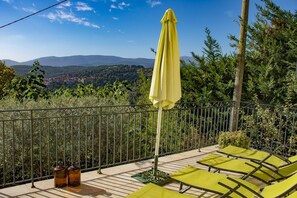 A terrace with a yellow umbrella, green lounge chairs, and a view of a town and mountains.