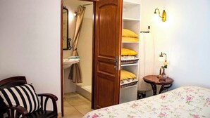 Une chambre à coucher comprenant un lit, une chaise, une armoire en bois, une étagère avec des coussins et une table de chevet avec une lampe.