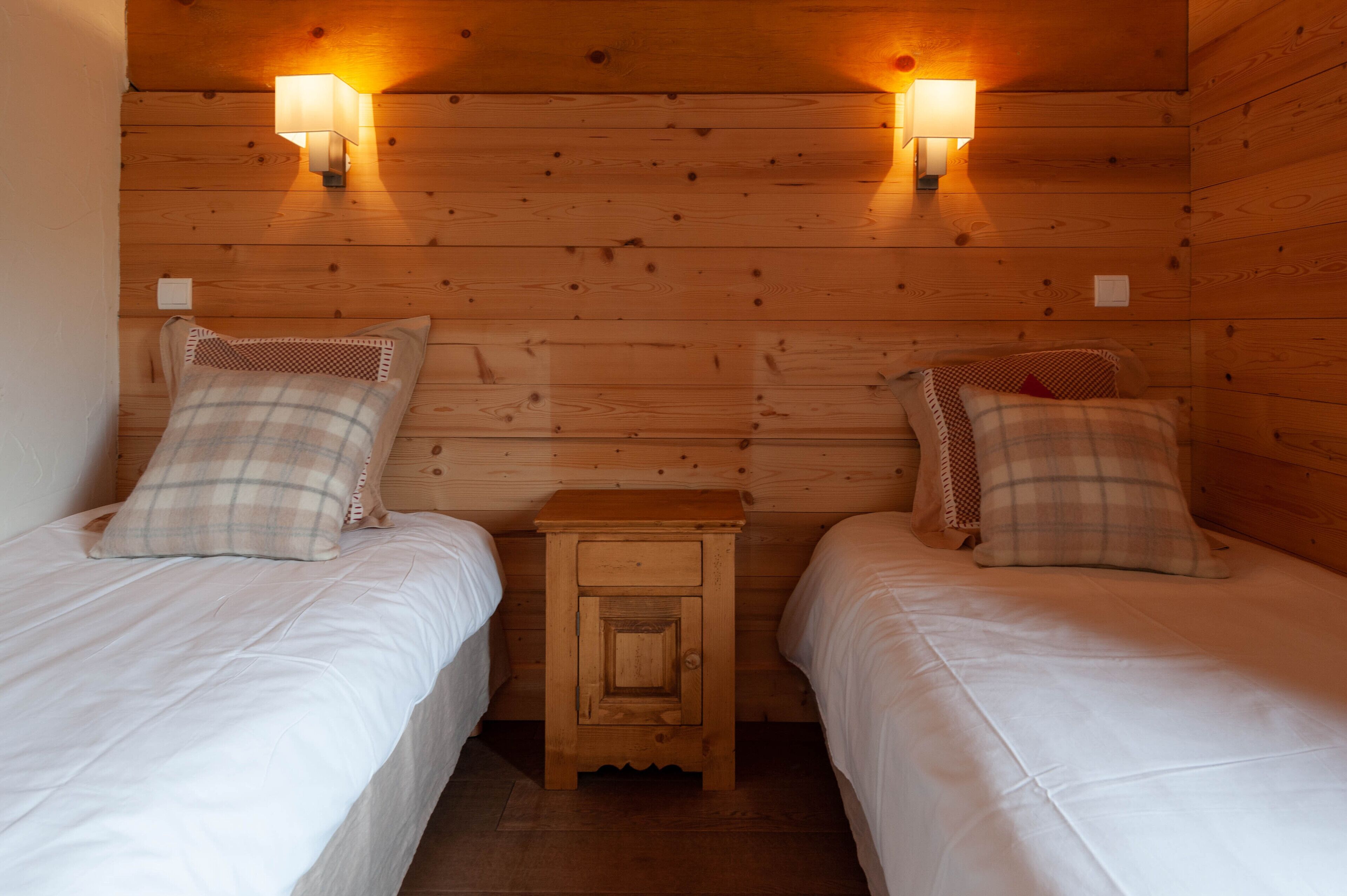 A wooden cabin room with two beds, each with a plaid pillow and a nightstand in between.