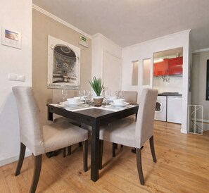 Un coin repas avec une table dressée pour quatre personnes, deux chaises de chaque côté, une petite plante en guise de décoration centrale et un tableau accroché au mur.