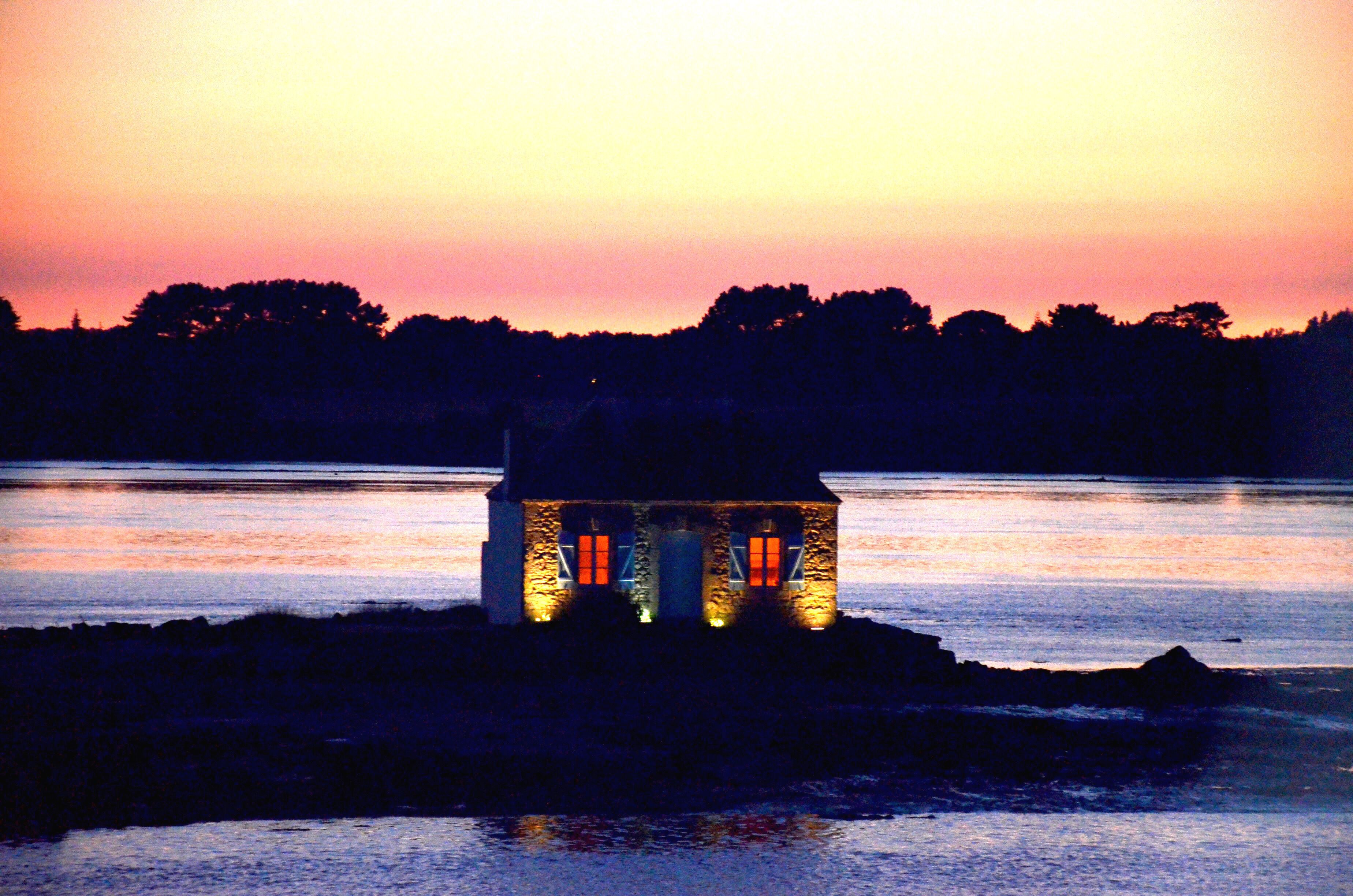 A small stone building with illuminated windows, situated on a lakeshore at sunset.