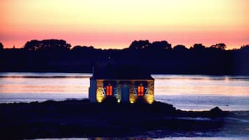 A small stone building with illuminated windows, situated on a lakeshore at sunset.