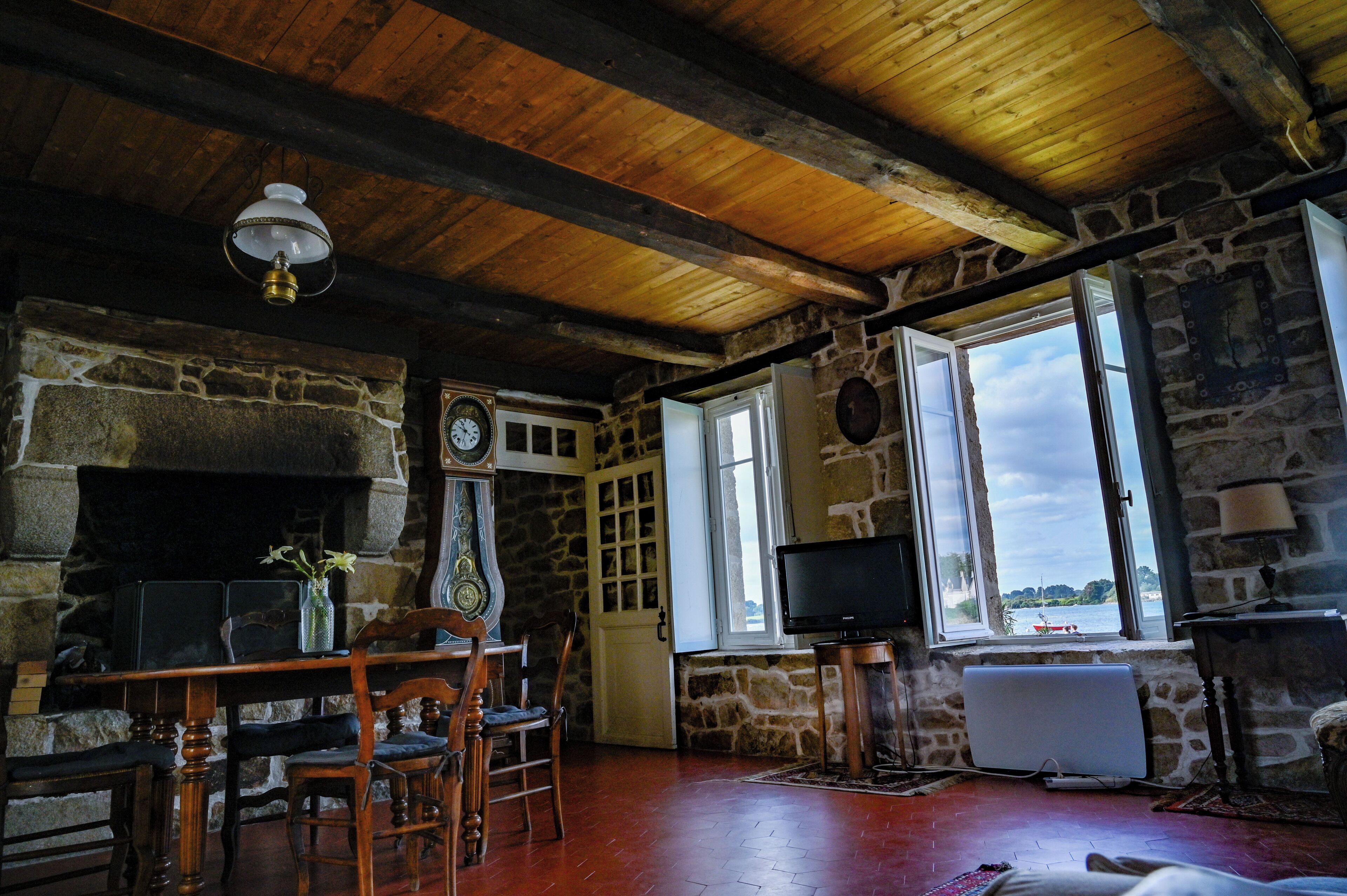 Quarto rústico com lareira de pedra, vigas de madeira no teto, uma mesa de madeira com cadeiras, televisão e vista para a água.