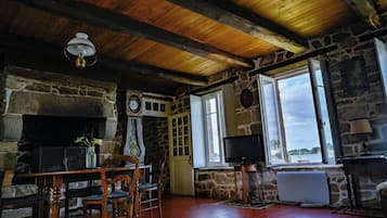 Quarto rústico com lareira de pedra, vigas de madeira no teto, uma mesa de madeira com cadeiras, televisão e vista para a água.