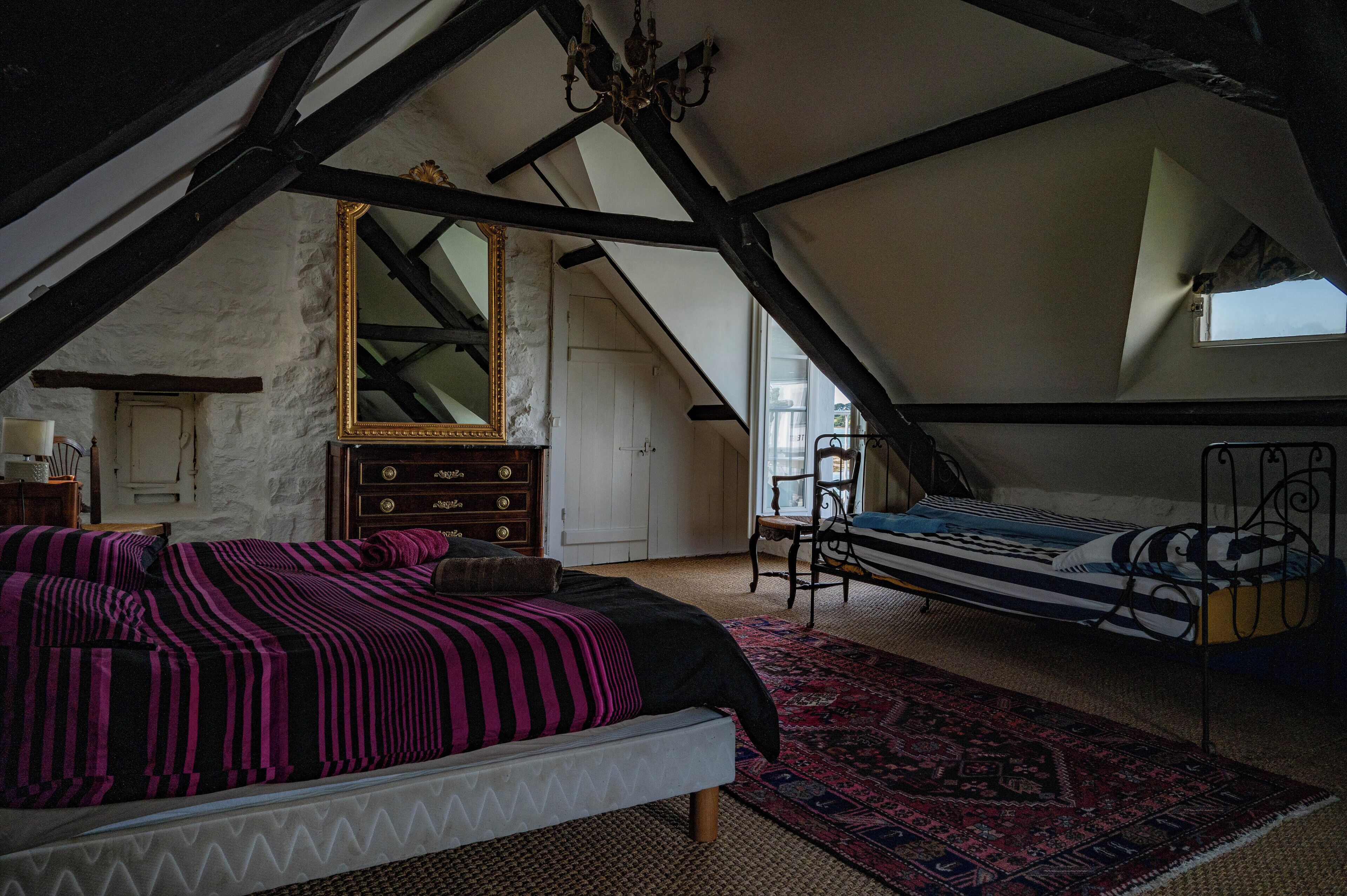 Ein Schlafzimmer mit schrägen Wänden, einem Bett mit gemustertem Überwurf, einer Kommode, einem Spiegel und einem Fenster.