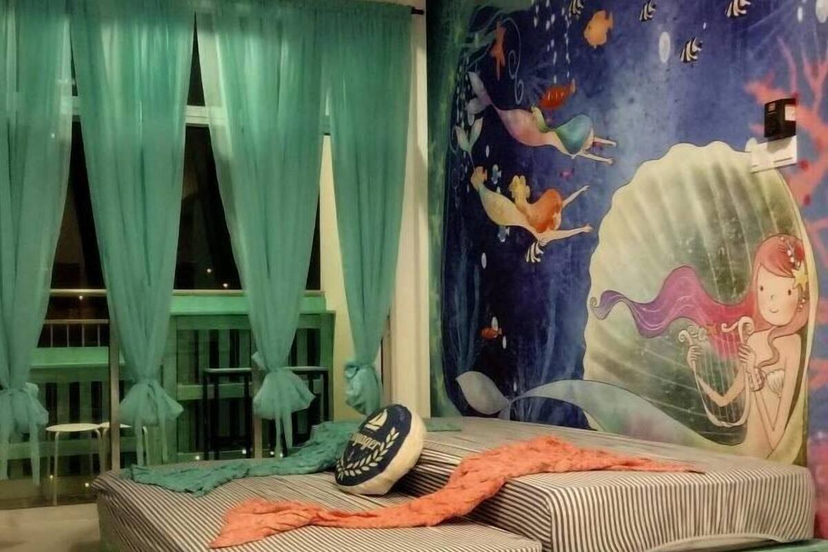 A room with a bed featuring striped bedding, a mural of a mermaid, teal curtains, and a small table with chairs.