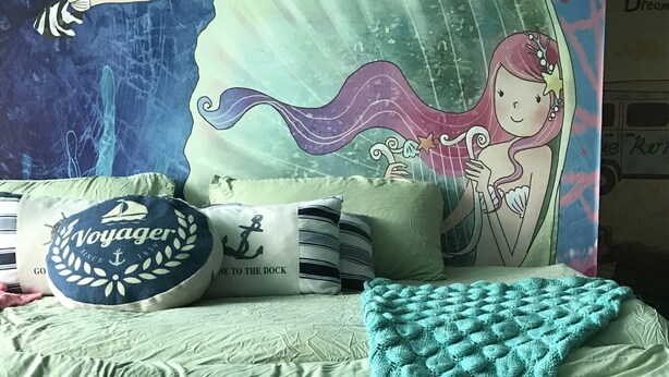 Ein Bett mit einem türkisfarbenen, gestrickten Meerjungfrauen-Schwanz, Kissen mit nautischen Motiven und ein Meerjungfrauen-Poster.