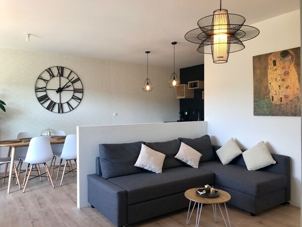 A modern living room with a dark grey sofa, a round coffee table, and a dining area with white chairs and a wooden table. A large wall clock and a painting are visible.