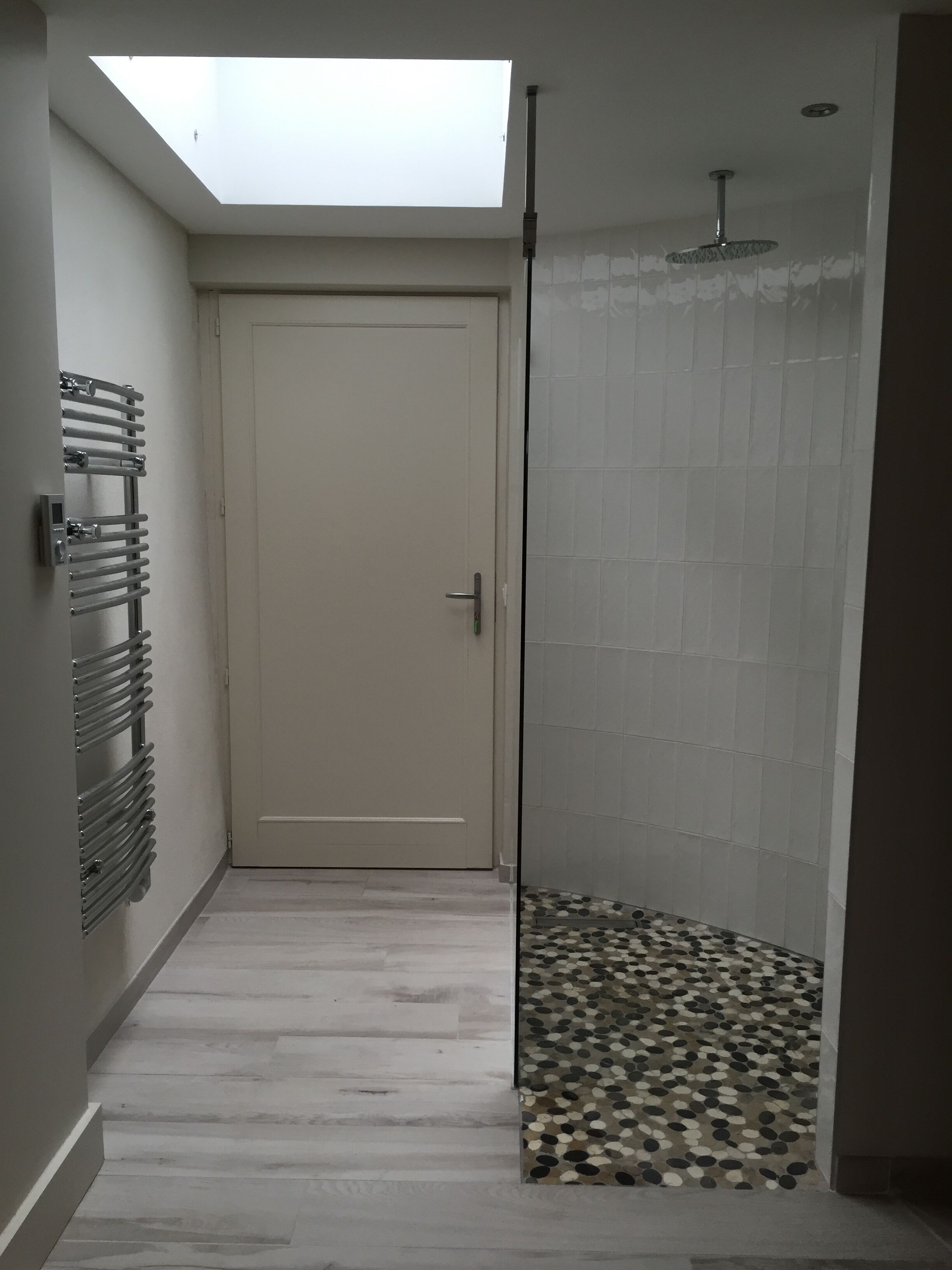 Un couloir étroit avec un puits de lumière, une porte, une douche avec un sol carrelé et un radiateur.