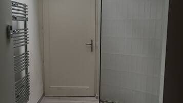 Un couloir étroit avec un puits de lumière, une porte, une douche avec un sol carrelé et un radiateur.
