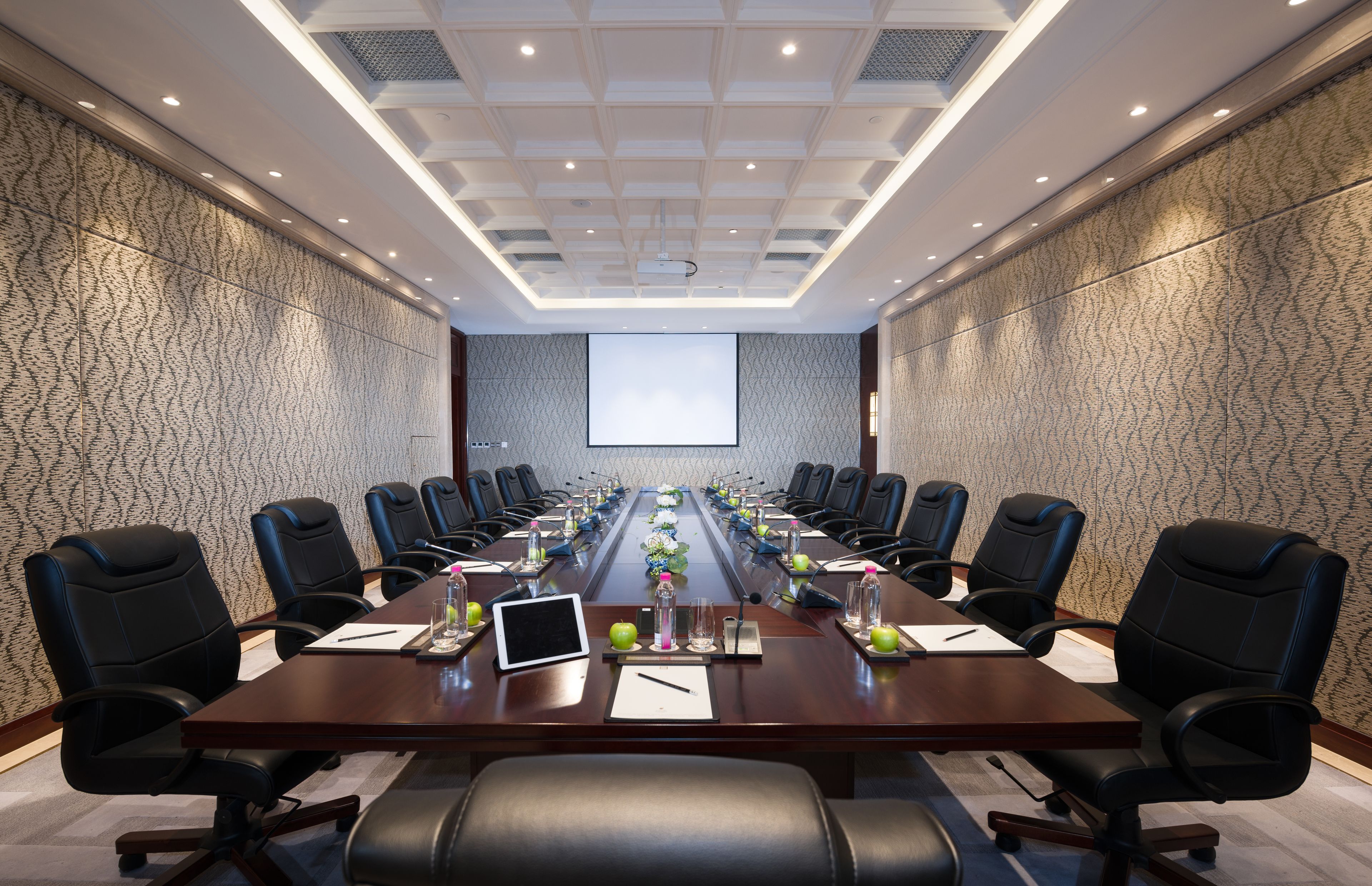 A conference room with a long table, black chairs, and a projector screen.