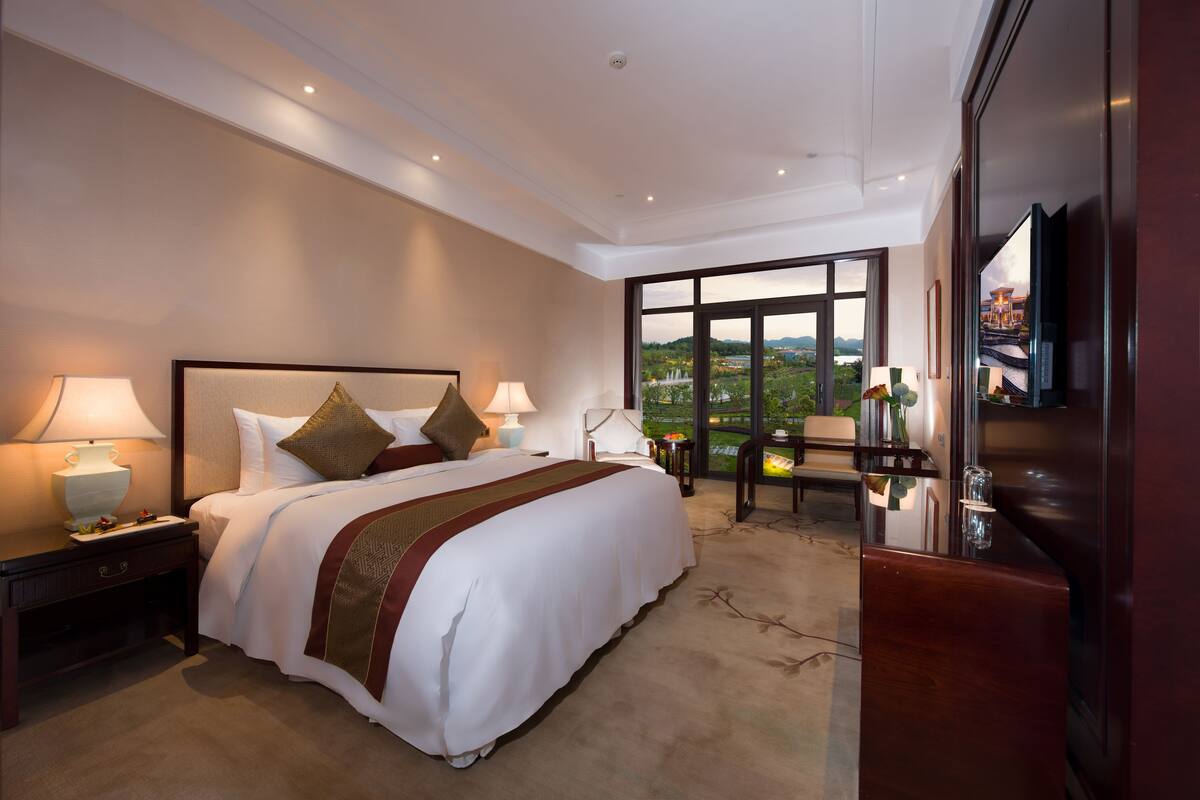 Hotelkamer met een groot bed, twee bedlampen, een nachtkastje, een televisie en uitzicht op een stadsbeeld door grote ramen.