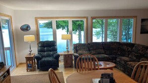Una sala de estar con un sillón de cuero, un sofá, una mesa de comedor con sillas y ventanas amplias con vista a árboles y un cuerpo de agua.