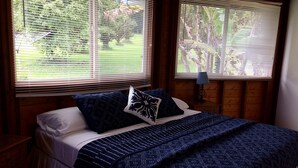 Un dormitorio con una cama, dos ventanas con persianas, una mesita de noche y una lámpara.