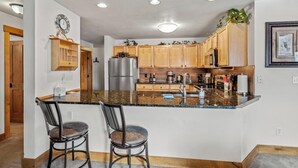 A modern kitchen with wooden cabinets, a stainless steel refrigerator, a granite countertop, and two bar stools. - Direct Unit Access,  Garage, 2 decks, Weber BBQ, Surround Sound Home Theater (Steamboat Springs)